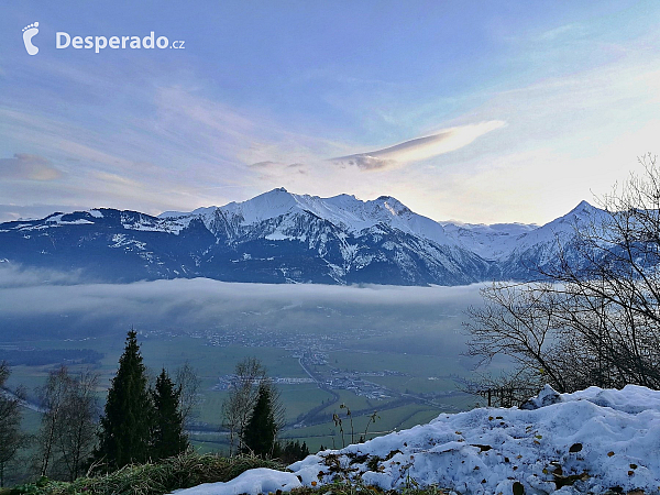 Slunce zapadající za zasněžené vrcholky hor a město Kaprun schovávající se pod nadýchanou mlhou.